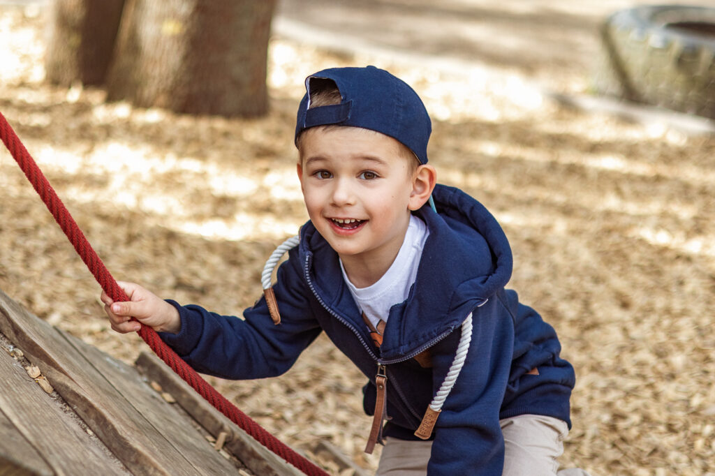 Auf der Schräge mit Seil klettert ein Junge mit blauem Cap und blauer Jacke hoch, Kindergartenfotografie im freien Spiel Berlin