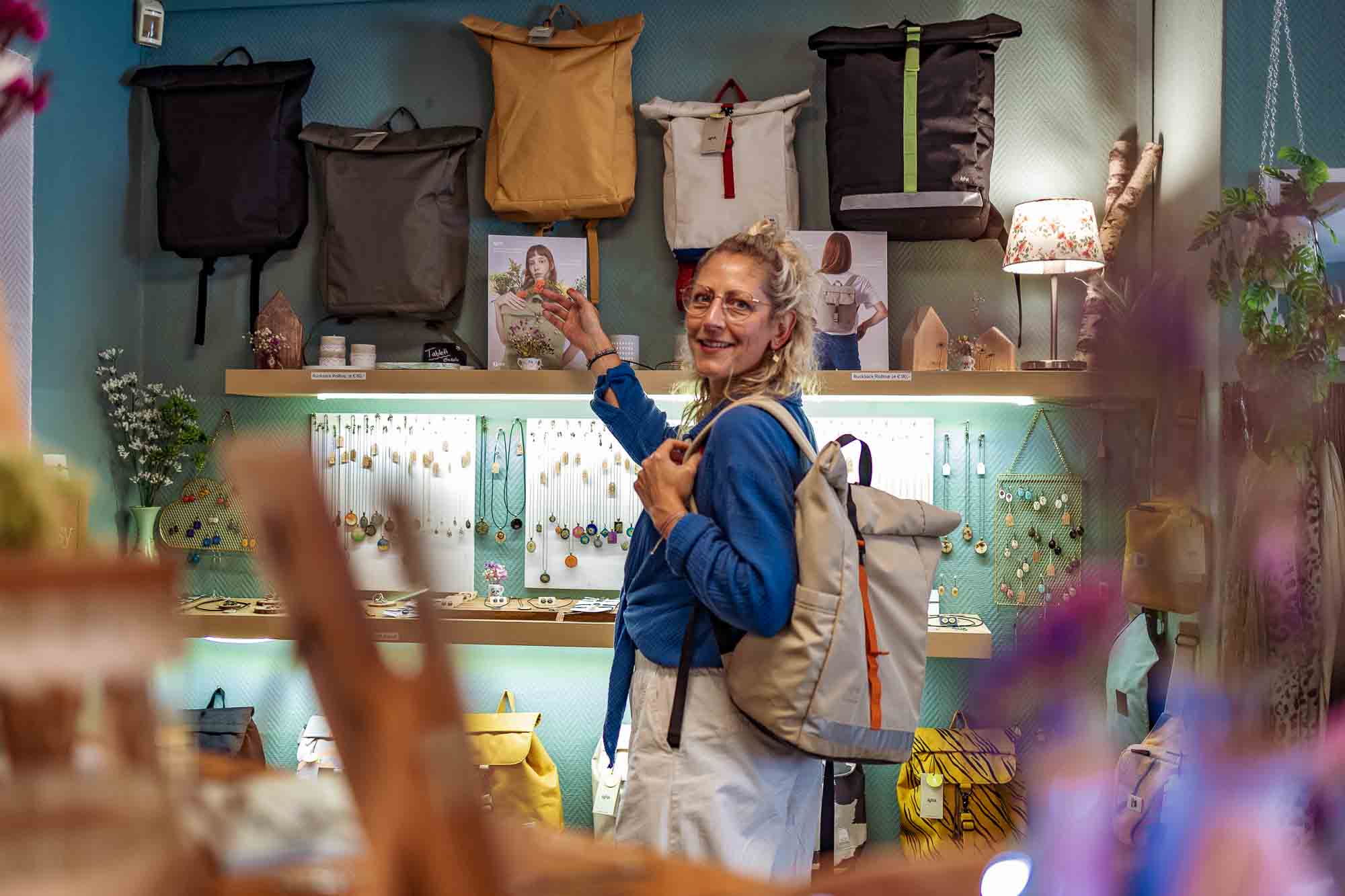 Unternehmensfotografie in Strausberg: Sunny, Inhaberin des Ladens Liebling Strausberg, präsentiert nachhaltige Rucksäcke aus recycelten Plastikflaschen, umgeben von handgefertigtem Schmuck aus Blüten und Blättern – authentische Businessfotografie mit Fokus auf kreative und umweltbewusste Produkte.